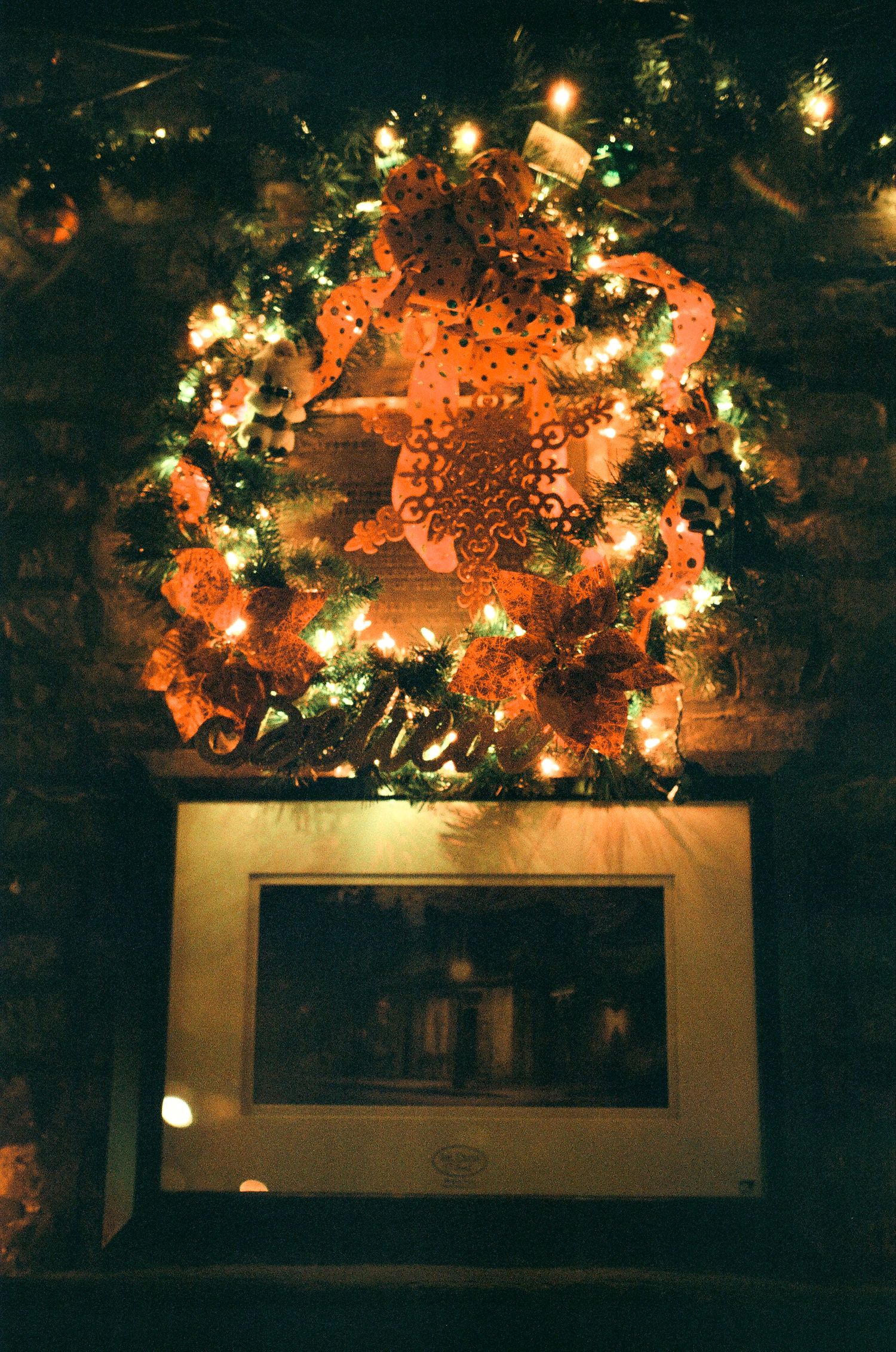 An illuminated holiday wreath with orange lights hangs above a framed display on a brick wall.