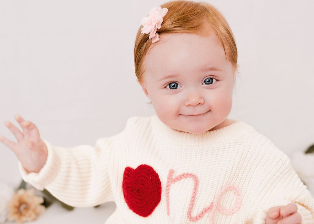 A baby wearing a cream sweater with a red heart design waves at the camera against a light background.