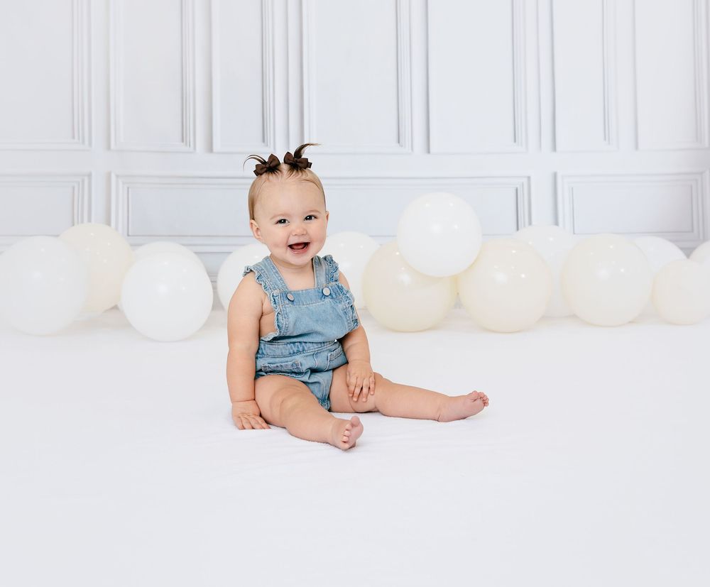 Happy baby in denim romper sitting on white floor surrounded by white balloons, smiling joyfully.