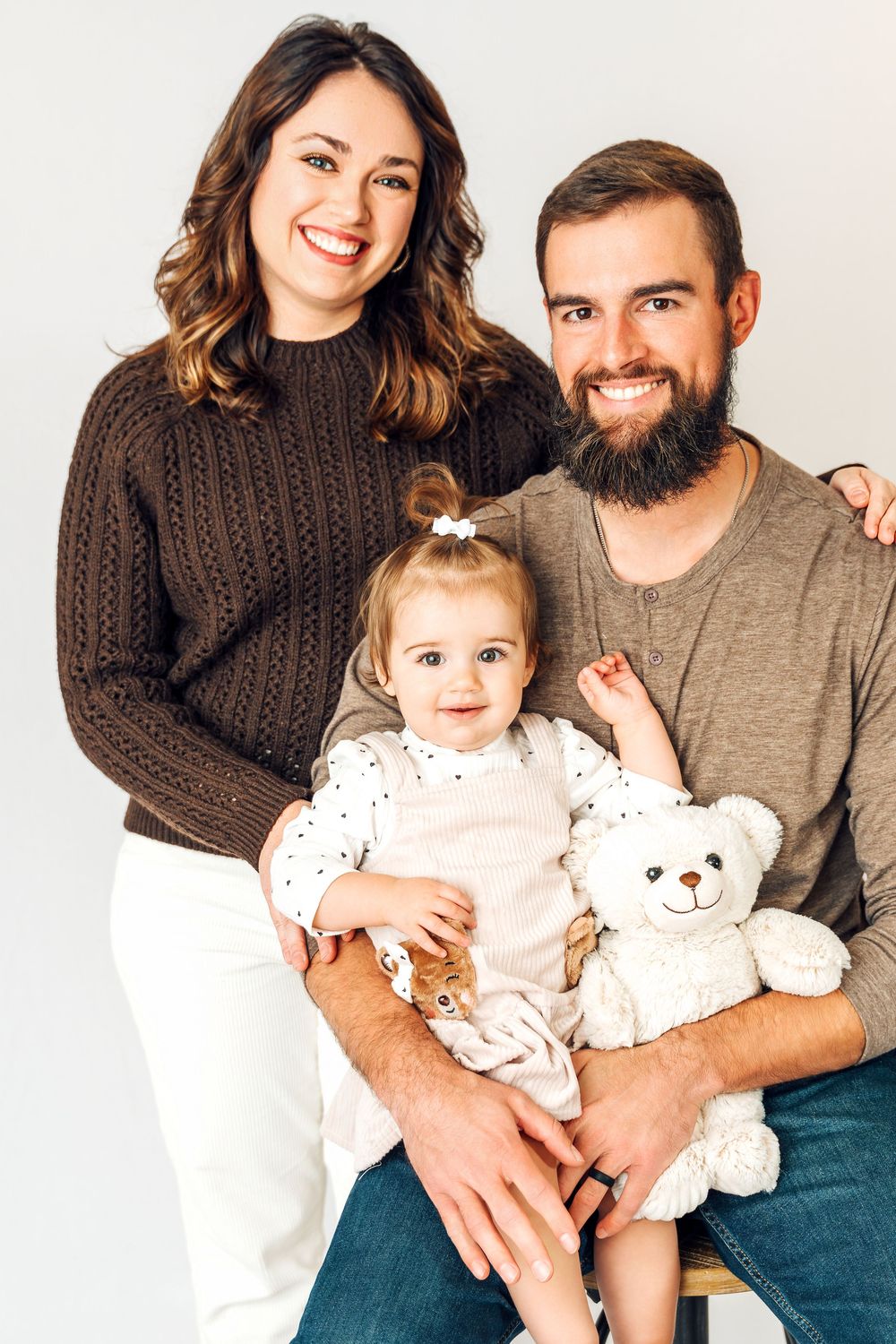 A family poses together for a portrait against a white background with their toddler holding a teddy bear.