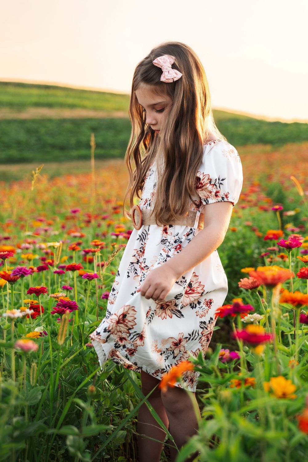 A white floral dress catches the summer breeze in a colorful field of orange and pink zinnias at sunset.