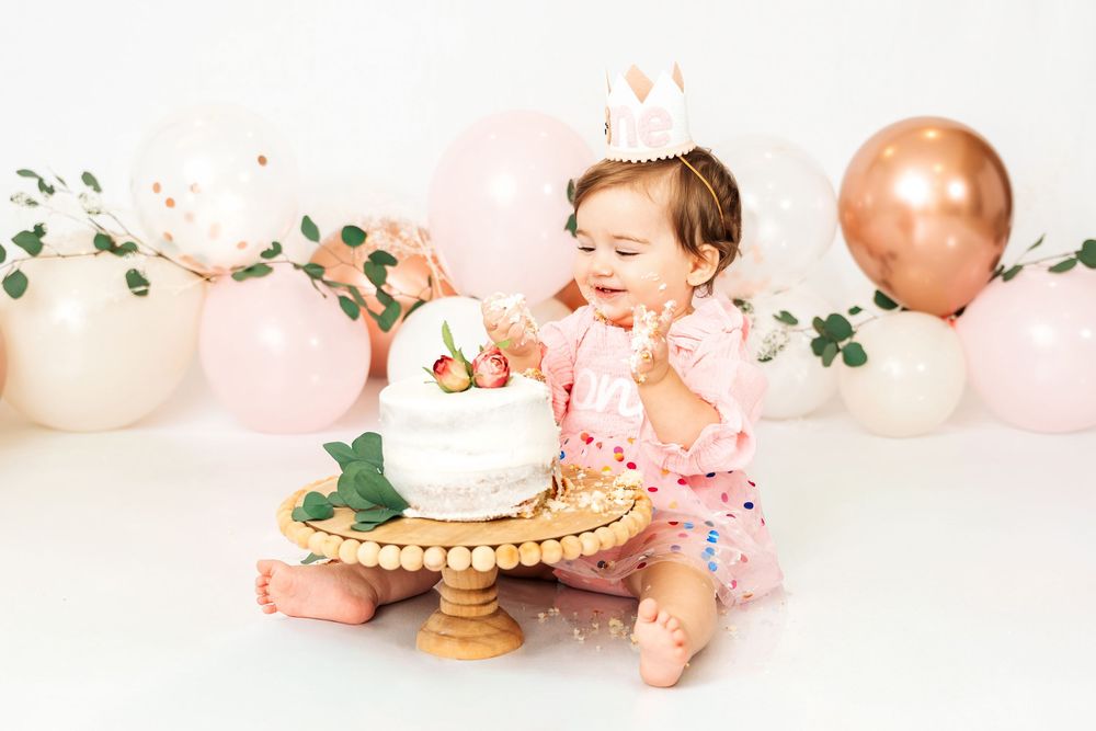 A baby sits in front of a pink and gold birthday cake setup with balloons and floral decorations.