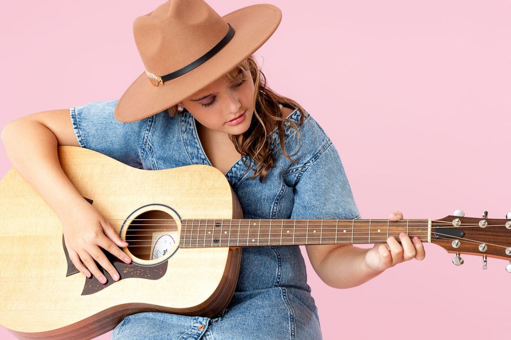 Young woman in denim dress and tan hat playing acoustic guitar against a pink background.