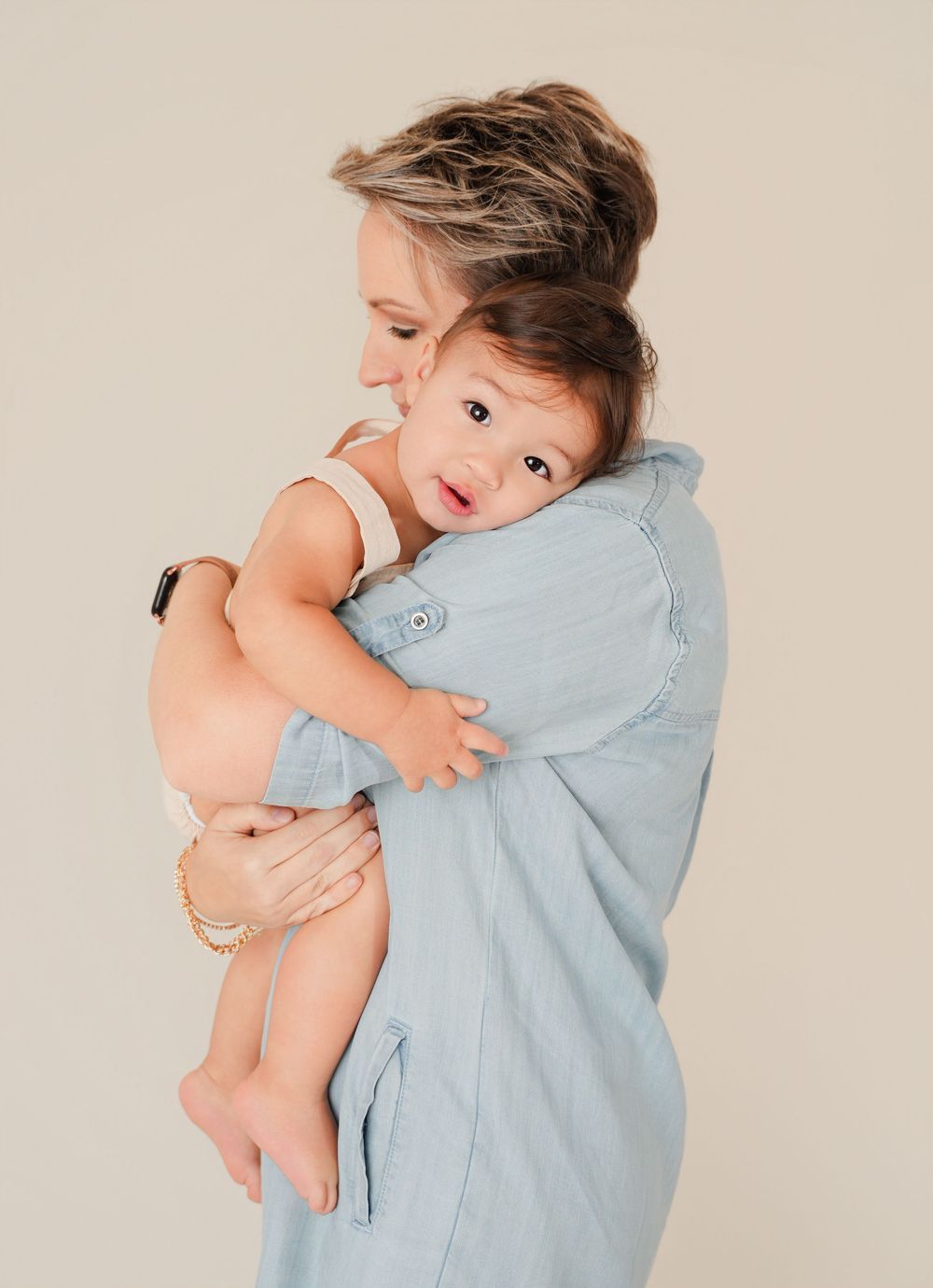 A woman in a light blue dress tenderly holds a baby against her shoulder in a soft, neutral-toned portrait.