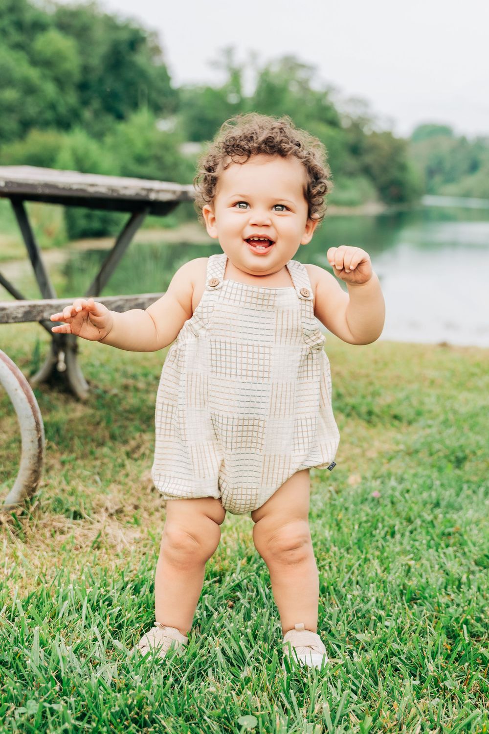 Happy curly-haired baby in beige romper stands on green grass near a pond outdoors.