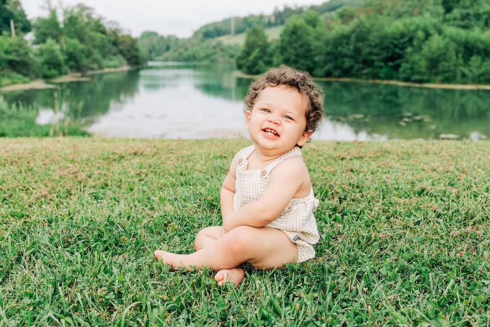 A happy toddler with curly hair sits barefoot on green grass near a peaceful lake, smiling brightly.