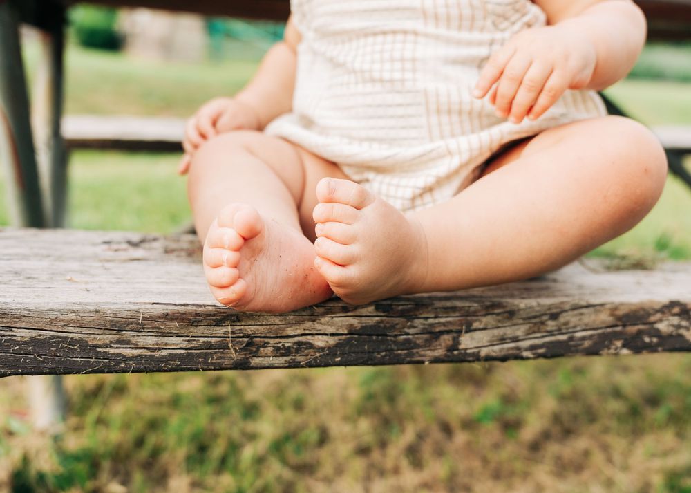 Close-up of adorable baby feet and legs sitting on a rustic wooden bench outdoors in nature.