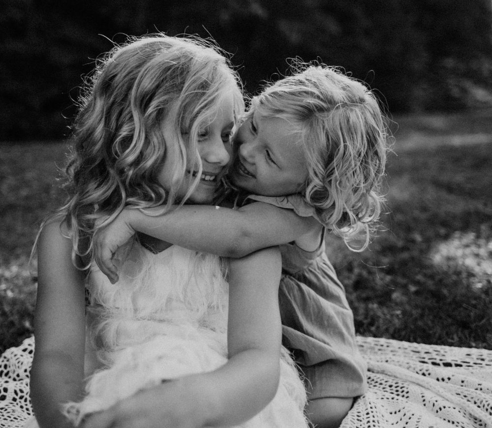 Two young children in white dresses share a tender moment outdoors in black and white.