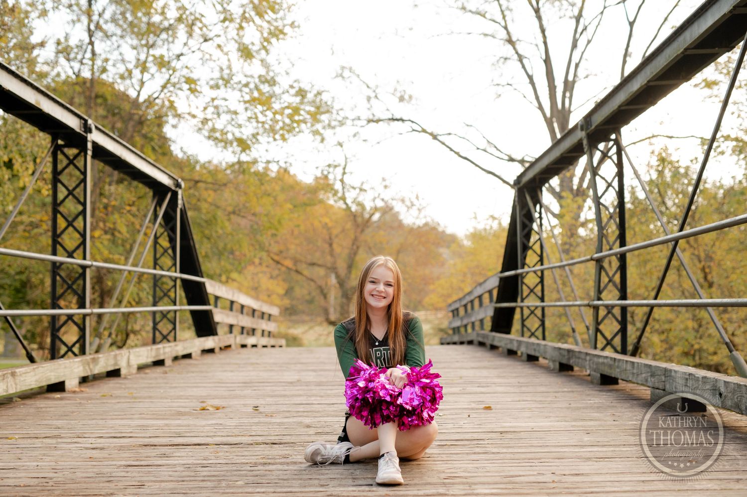 Cheerleader with pink pom poms crouches on wooden bridge during fall photo session.