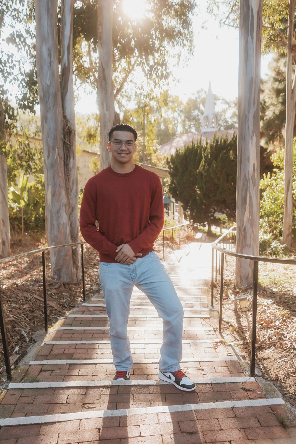 A person in a red sweater and light jeans poses on brick steps with metal railings.