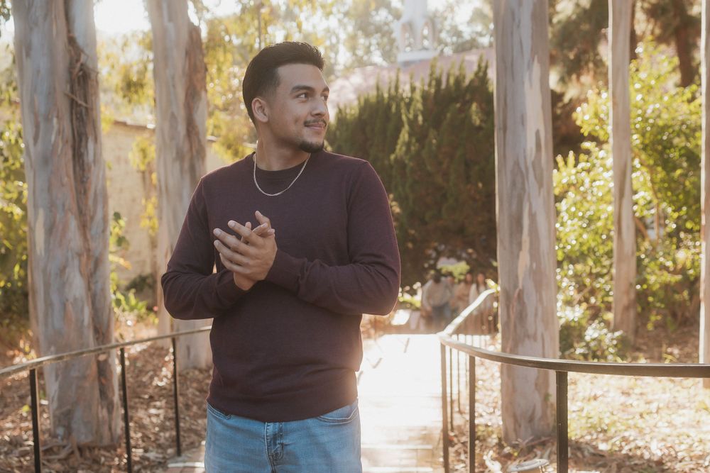 A person in a brown sweater stands thoughtfully on an outdoor path with sunlit trees.