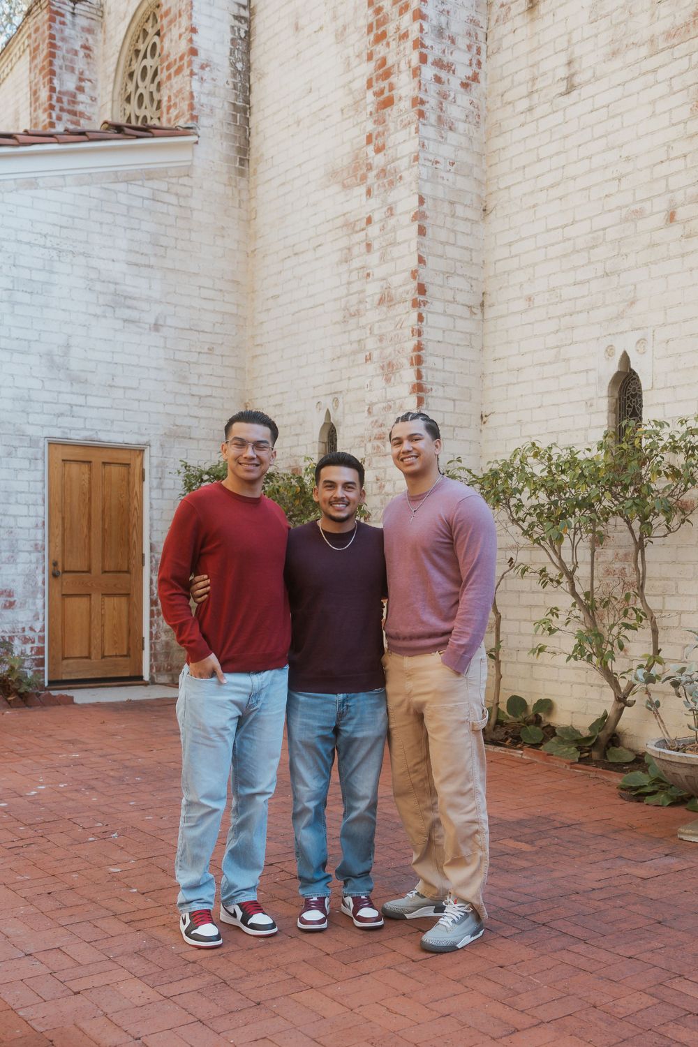 Three people pose together in front of a white brick wall with a wooden door.