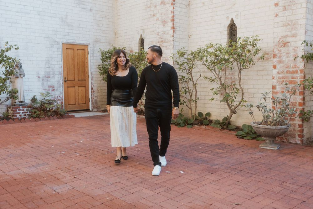 A couple walks together on red brick pavement in front of a white building wall.
