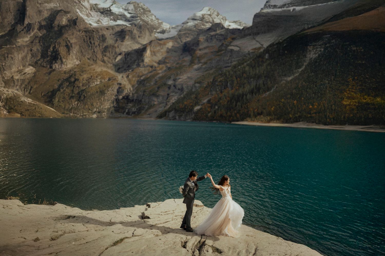 Wedding couple stands on rocky outcrop overlooking turquoise mountain lake.