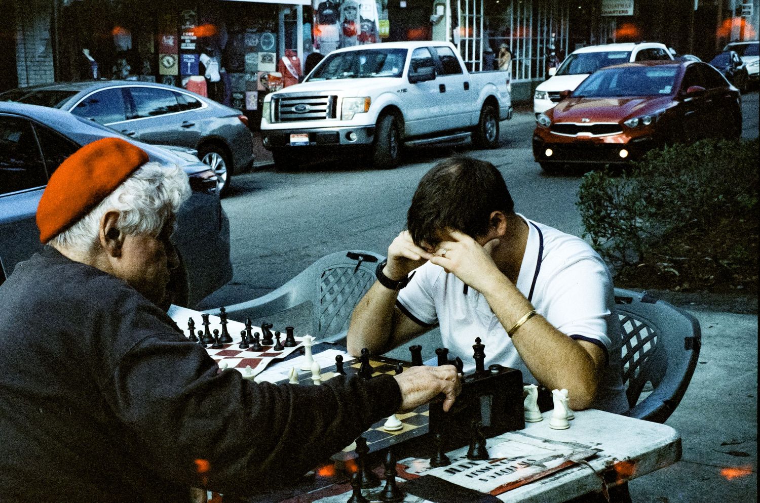 Two people engaged in a chess game on an outdoor table with cars in the background.