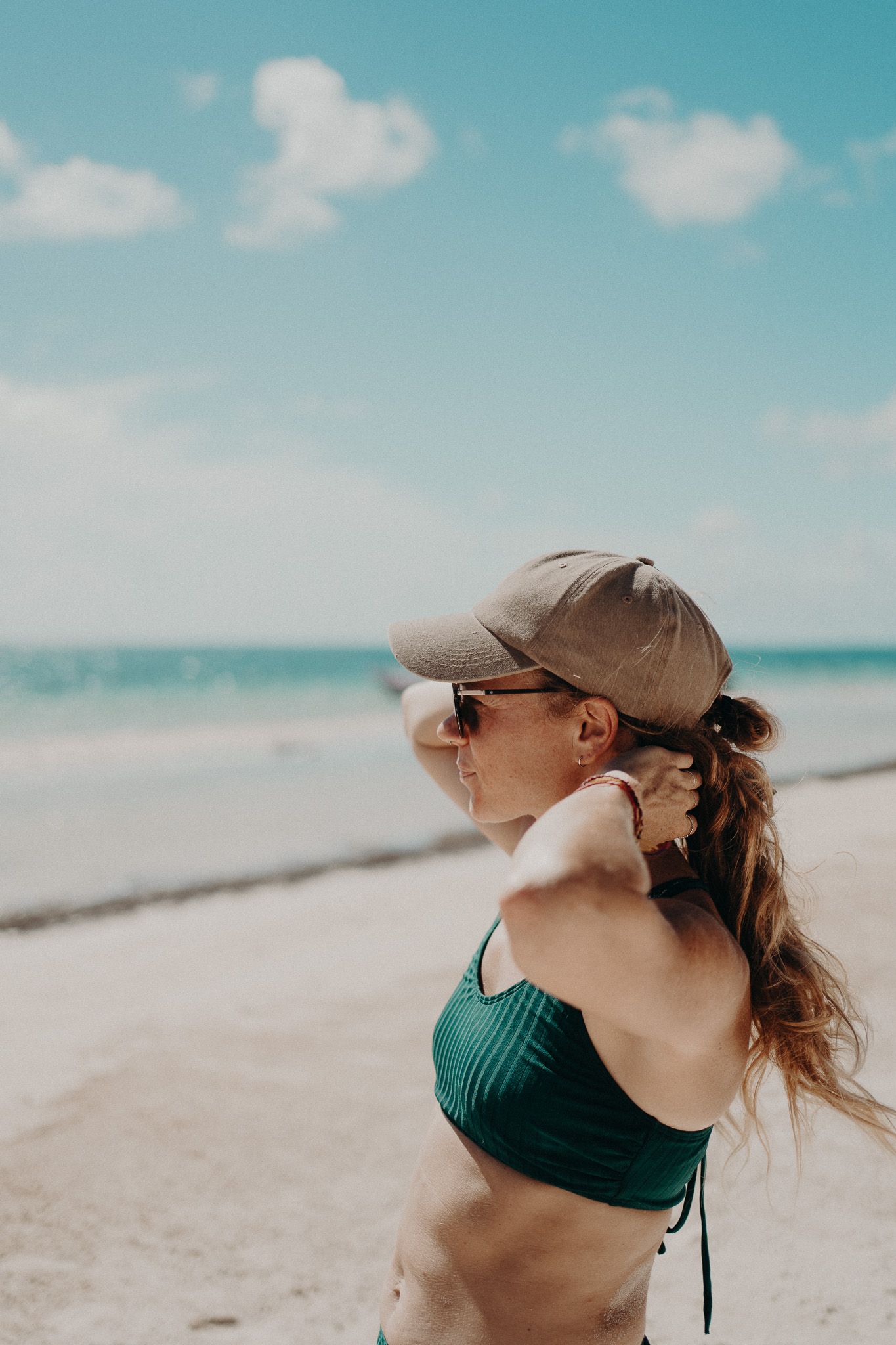 Woman in teal swimsuit and baseball cap enjoys ocean breeze on sunny beach photoshoot in Puerto Morelos.