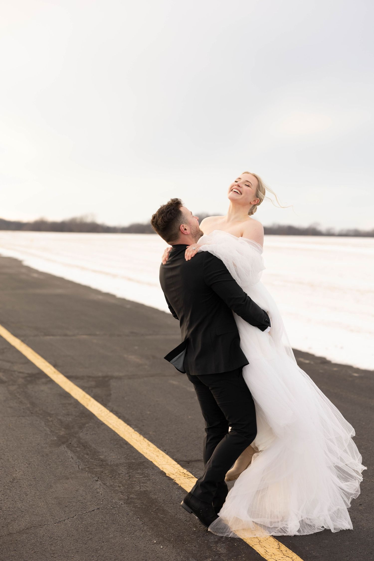 A couple in wedding attire embrace joyfully on an empty runway with snowy surroundings and overcast skies.
