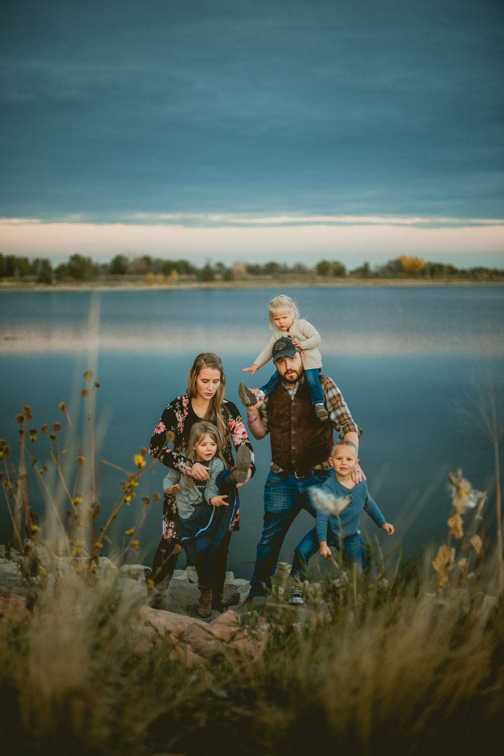 Family enjoys playful outdoor moment together by serene lake with tall grass in foreground at dusk.