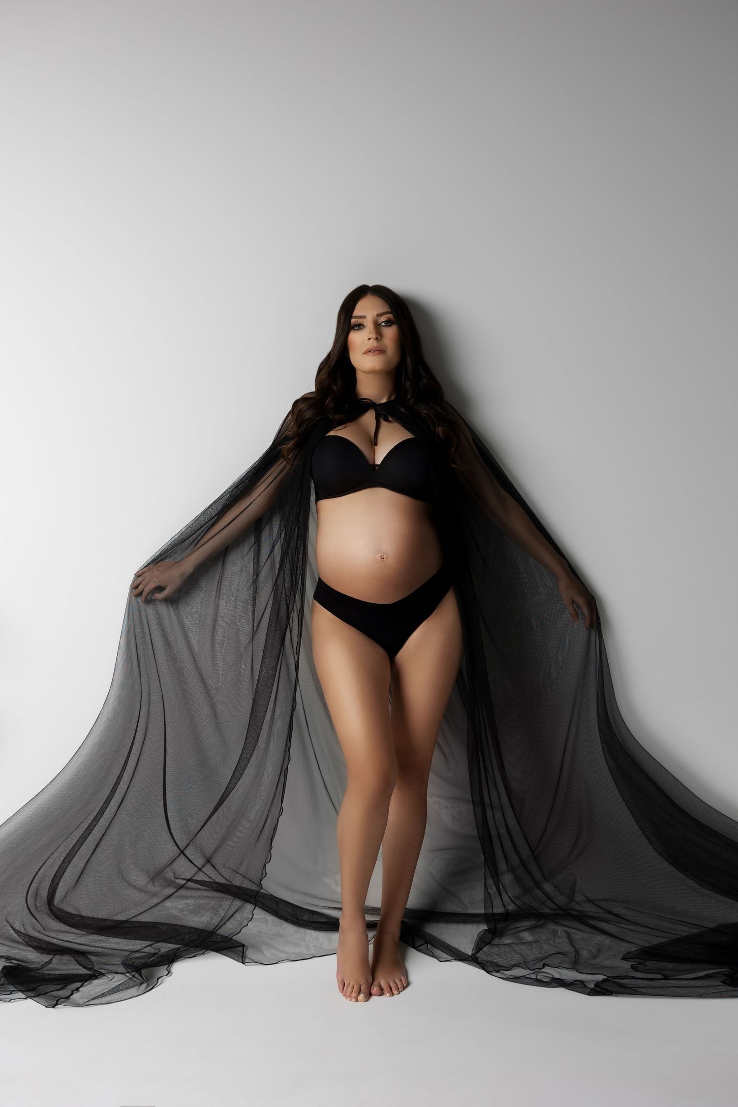 A person in black lingerie poses against white background while wearing flowing sheer black cape.