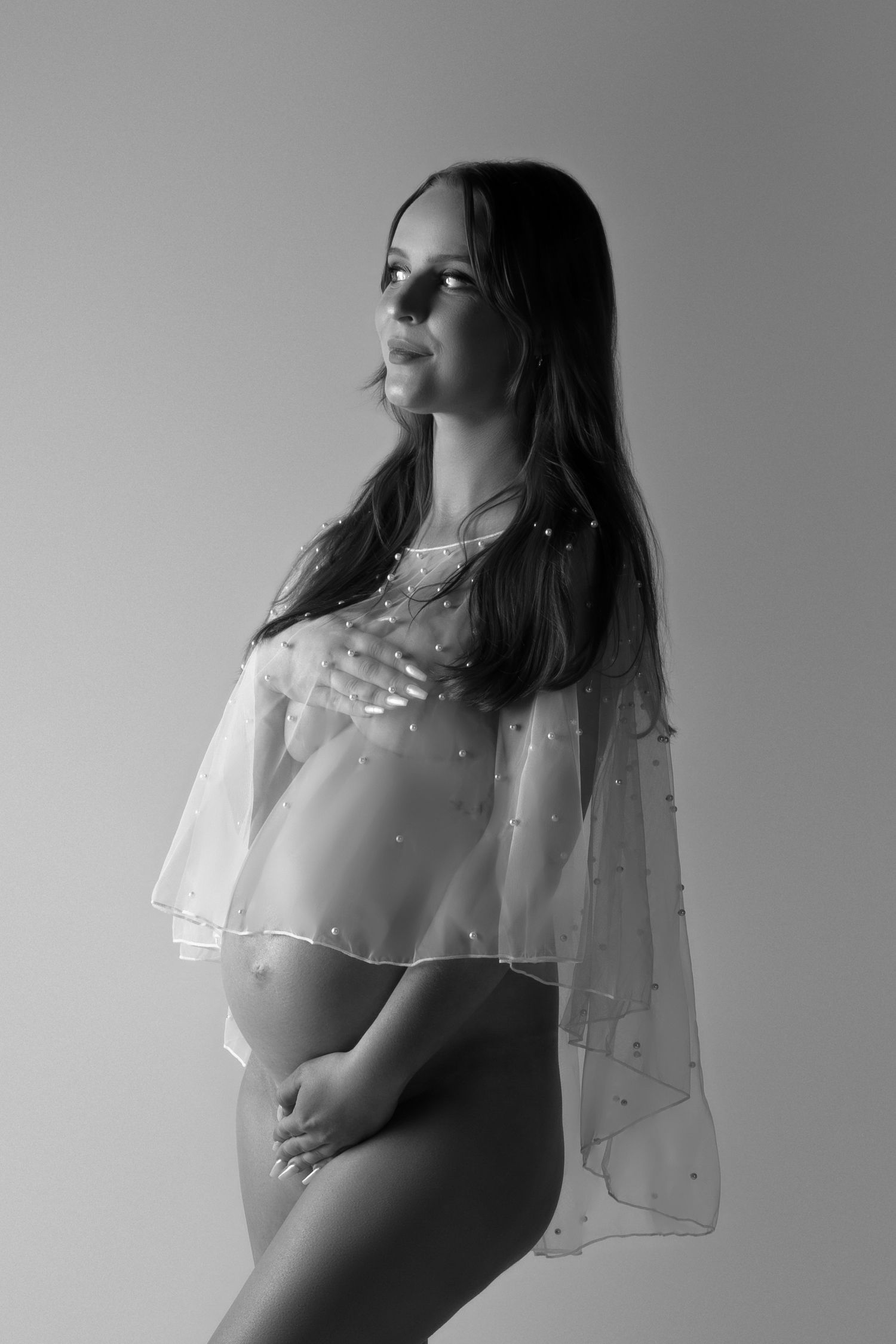 Black and white artistic maternity photo with sheer flowing veil and profile pose against plain backdrop.