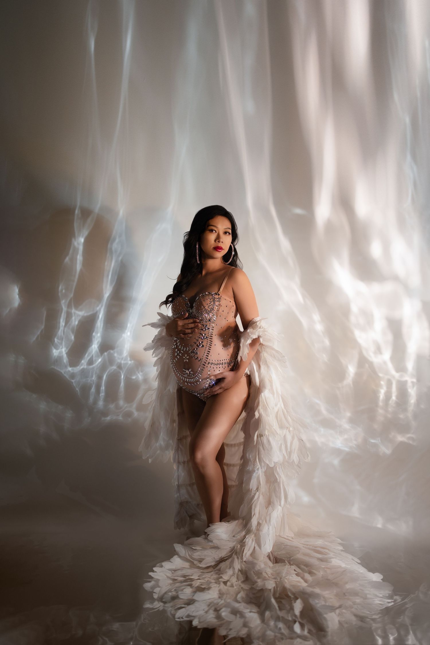 A figure poses dramatically in a sheer embellished bodysuit with flowing white fabric against an ethereal backlit backdrop.