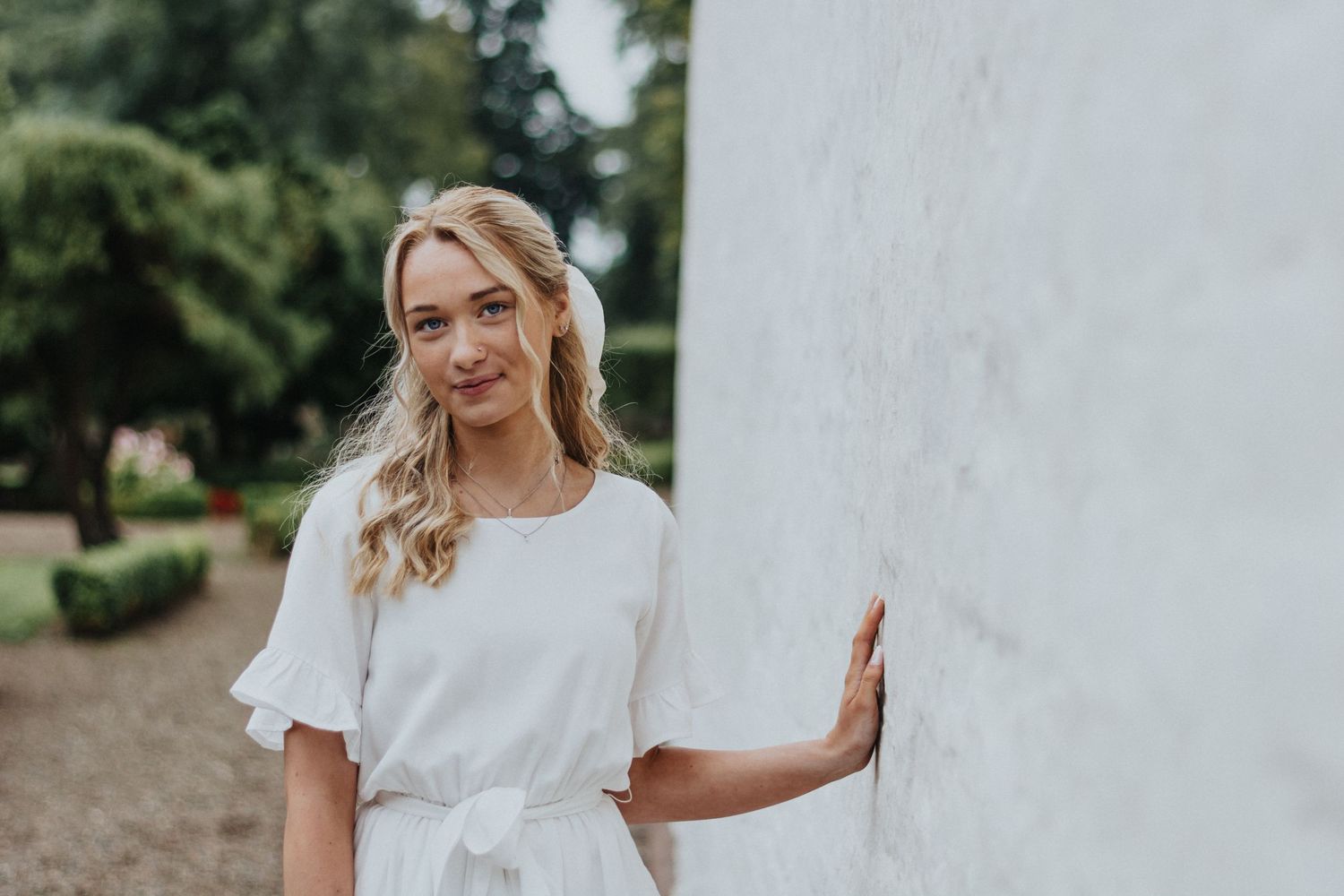 Woman in white dress poses against white wall in outdoor garden setting.