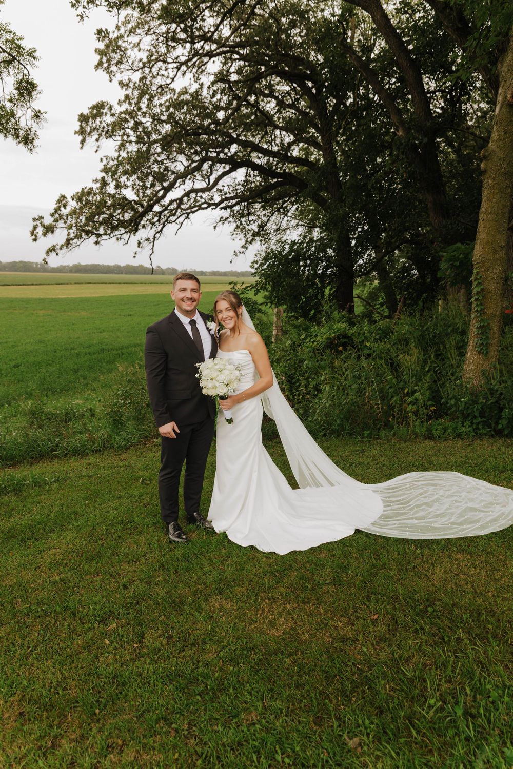 A wedding couple poses in a scenic outdoor setting with a flowing white veil caught in the breeze.