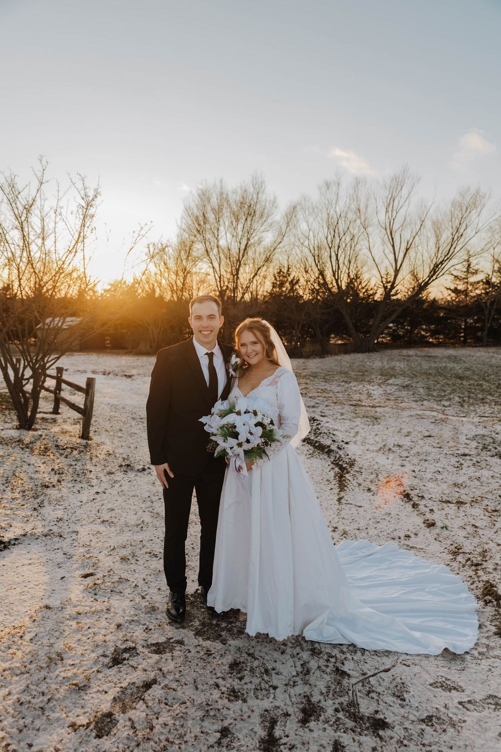 A couple poses in formal wedding attire on snowy ground at sunset with bare winter trees in the background.