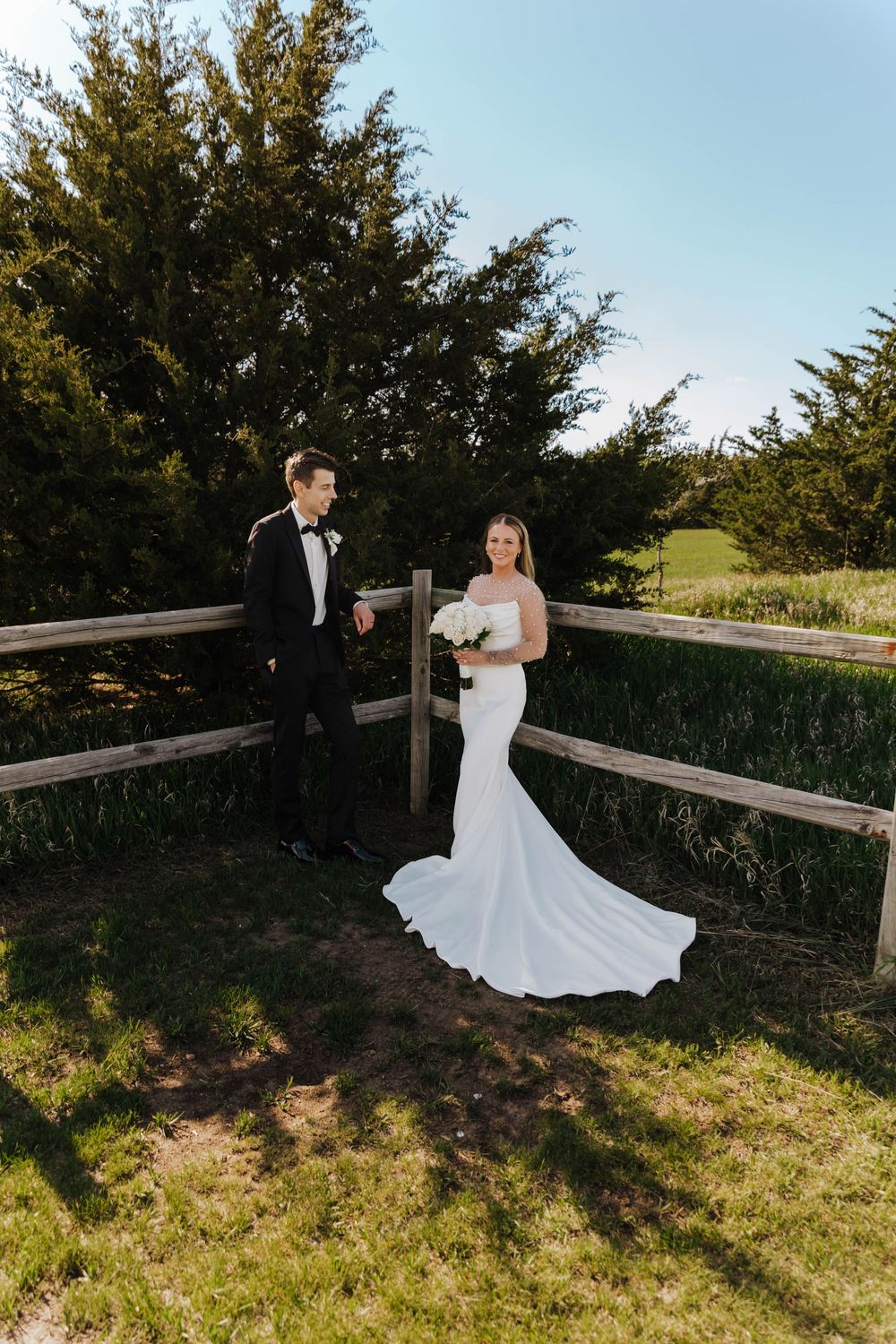 A couple poses for romantic wedding photos along a rustic wooden fence in golden afternoon light.
