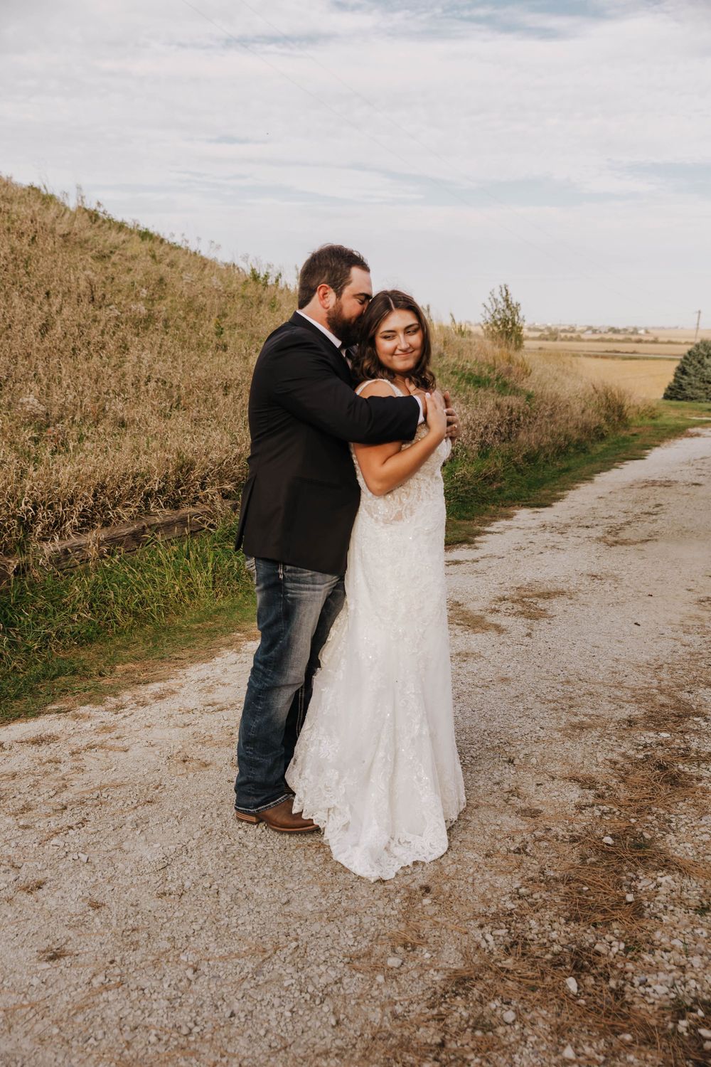 A couple shares an intimate moment on a dirt path surrounded by tall grass and open fields.