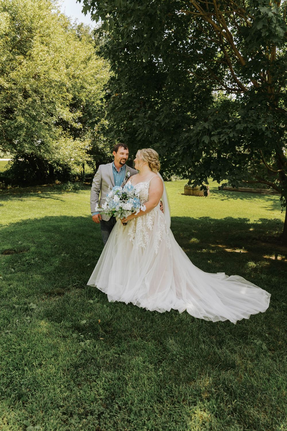 Newlywed couple shares romantic moments in a sun-dappled garden setting with lush green grass and trees.