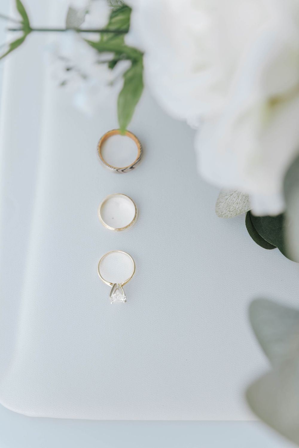 Three elegant wedding rings displayed on a white surface with delicate white flowers and green foliage in soft focus.
