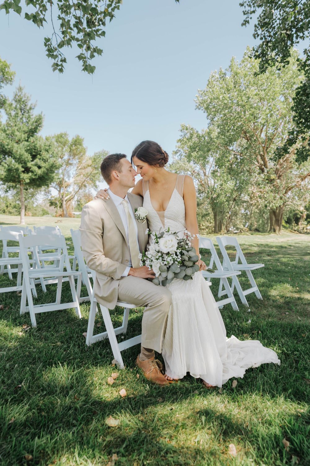 A newlywed couple shares tender moments on white chairs during their outdoor garden ceremony.