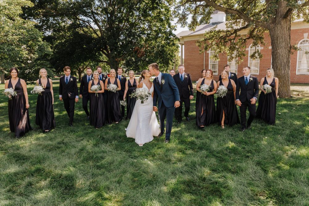 Wedding party in black and navy attire poses together on grassy lawn outside brick building.
