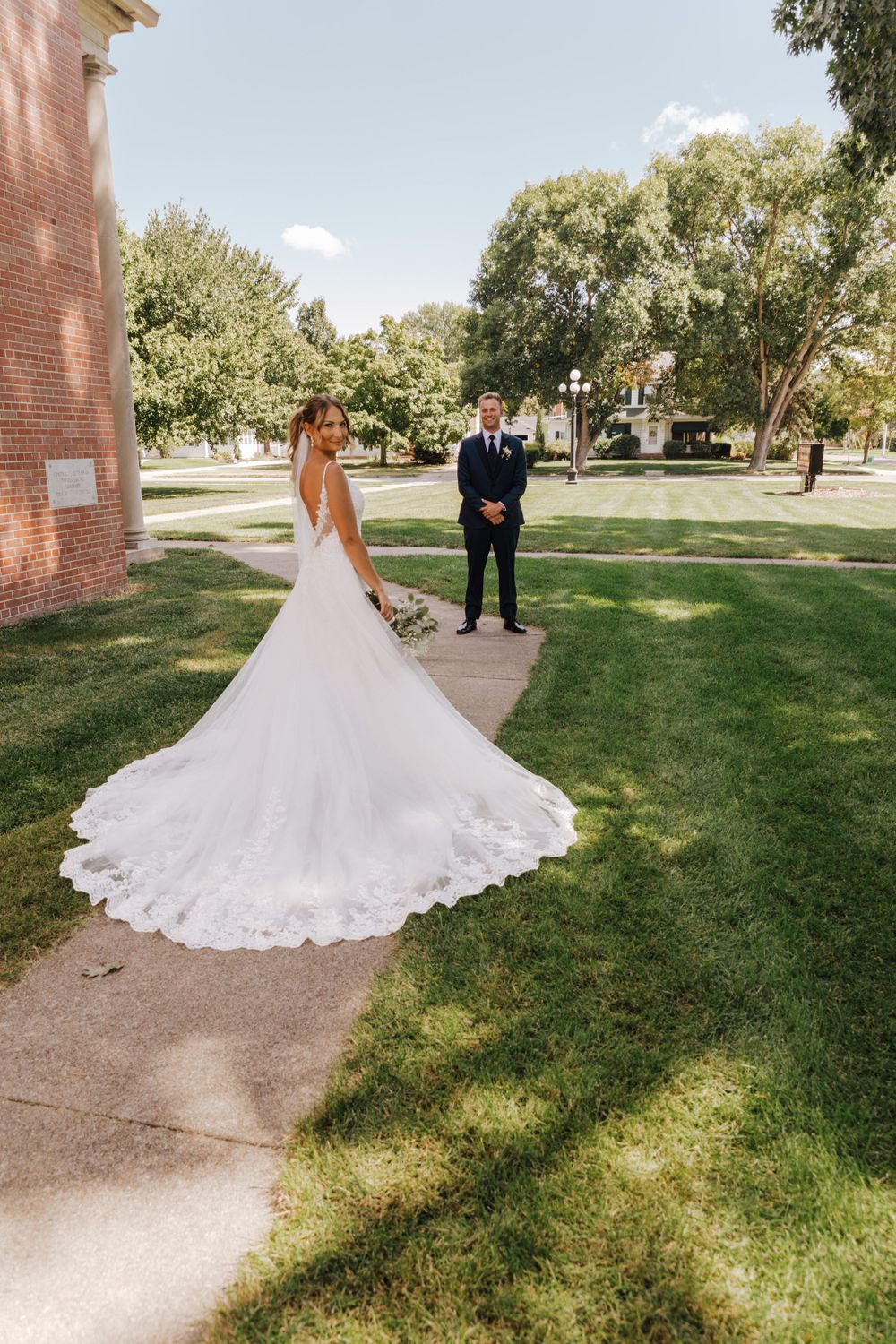 Bride in flowing lace train wedding dress stands on pathway with groom near brick building on sunny day.