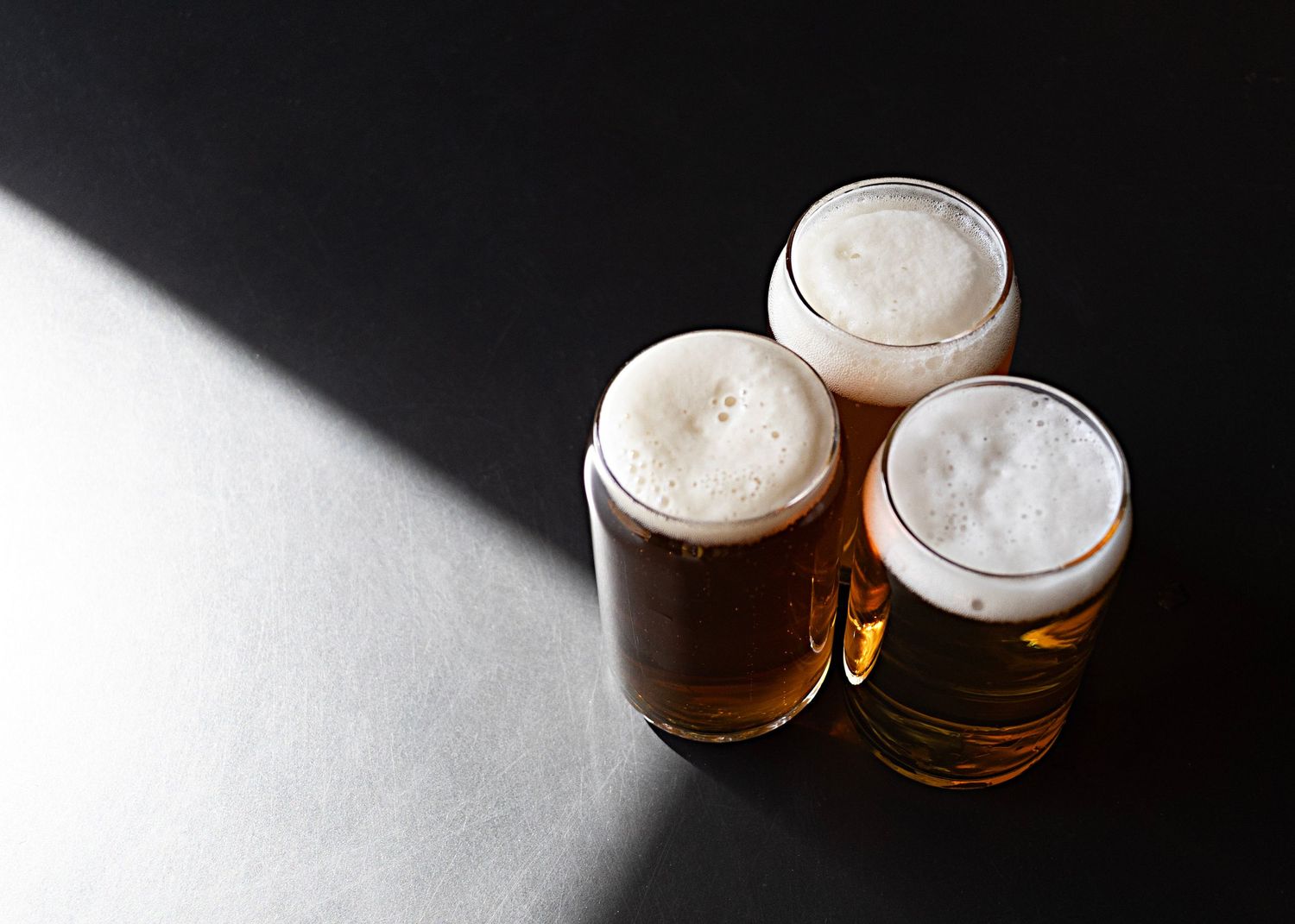 Three glasses of beer with frothy heads photographed against dark background with dramatic lighting.