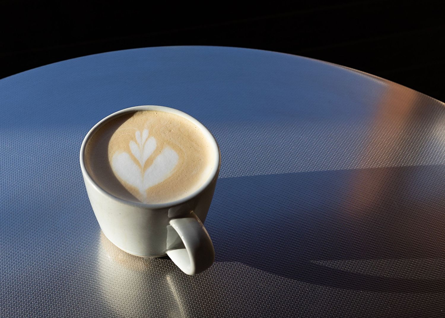 A white ceramic coffee cup with latte art featuring a delicate leaf design on a textured blue surface.