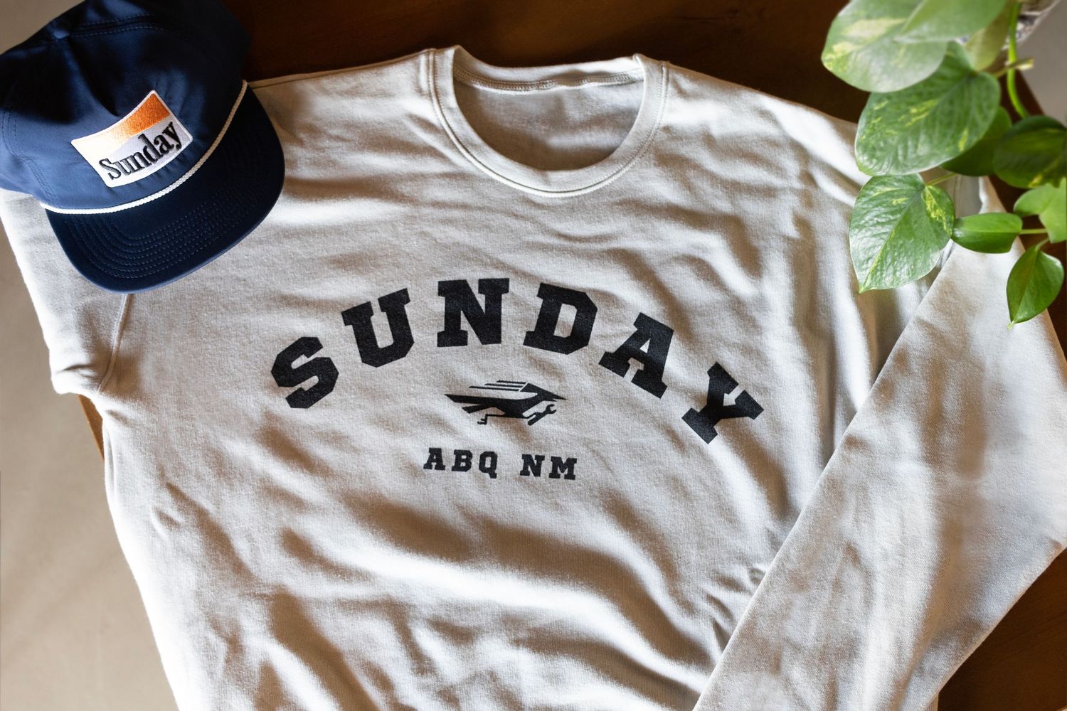 A grey t-shirt with SUNDAY text branding displayed next to a baseball cap and houseplant.