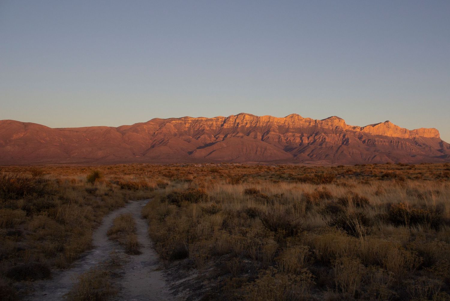 Dirt trail winding through desert landscape with red mountains glowing at sunset.
