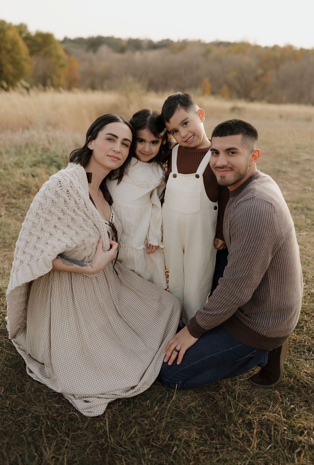 A family in neutral colored sweaters sits together on autumn grass during a golden hour outdoor photo session.