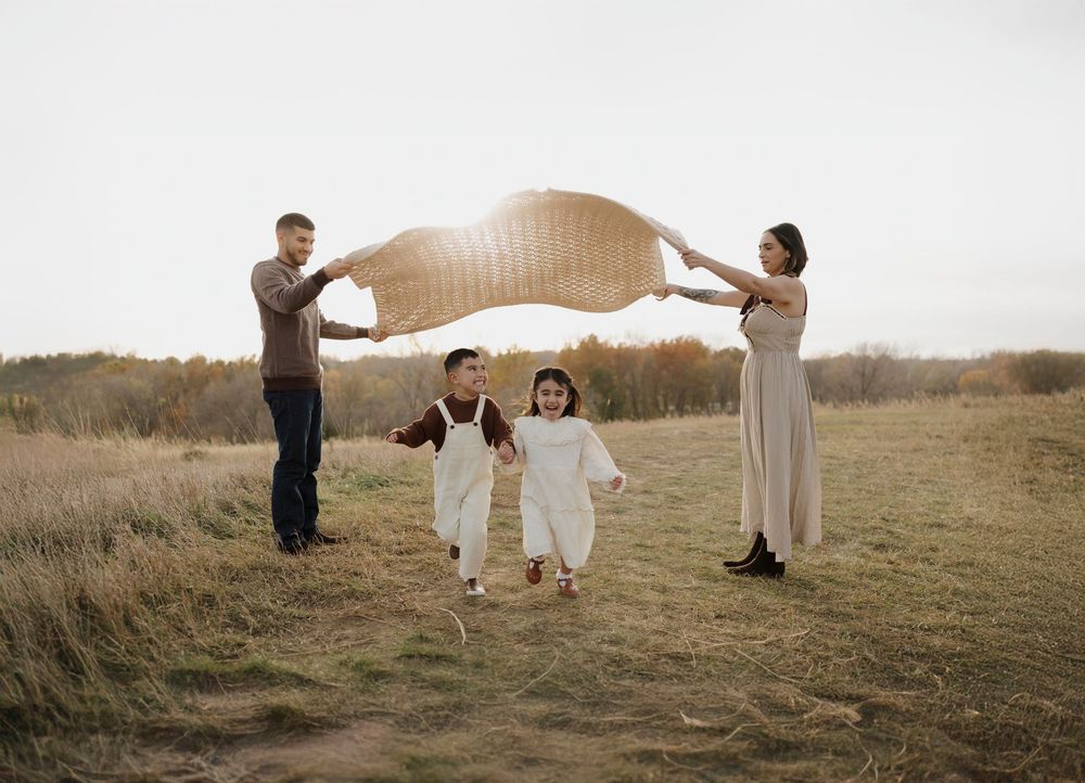 A family plays together in a golden field at sunset as parents hold up a blanket while children run underneath.