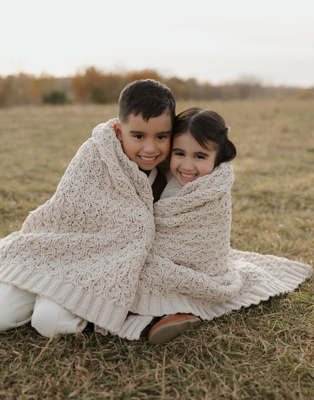 Two children wrapped in a cozy cream knit blanket lying in a grassy field at sunset.
