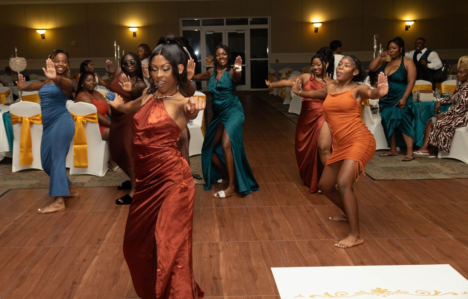Bridesmaids in colorful satin gowns dancing energetically on the dance floor at a wedding reception.