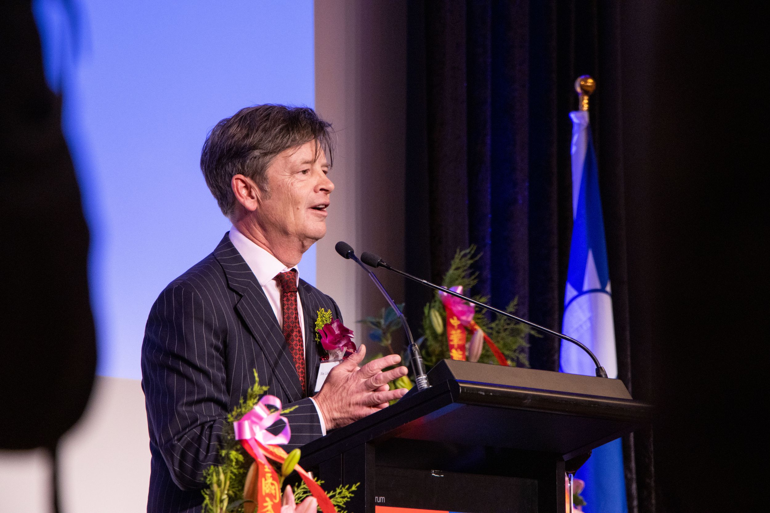 Speaker delivers remarks at podium with floral decorations and flags during formal event.