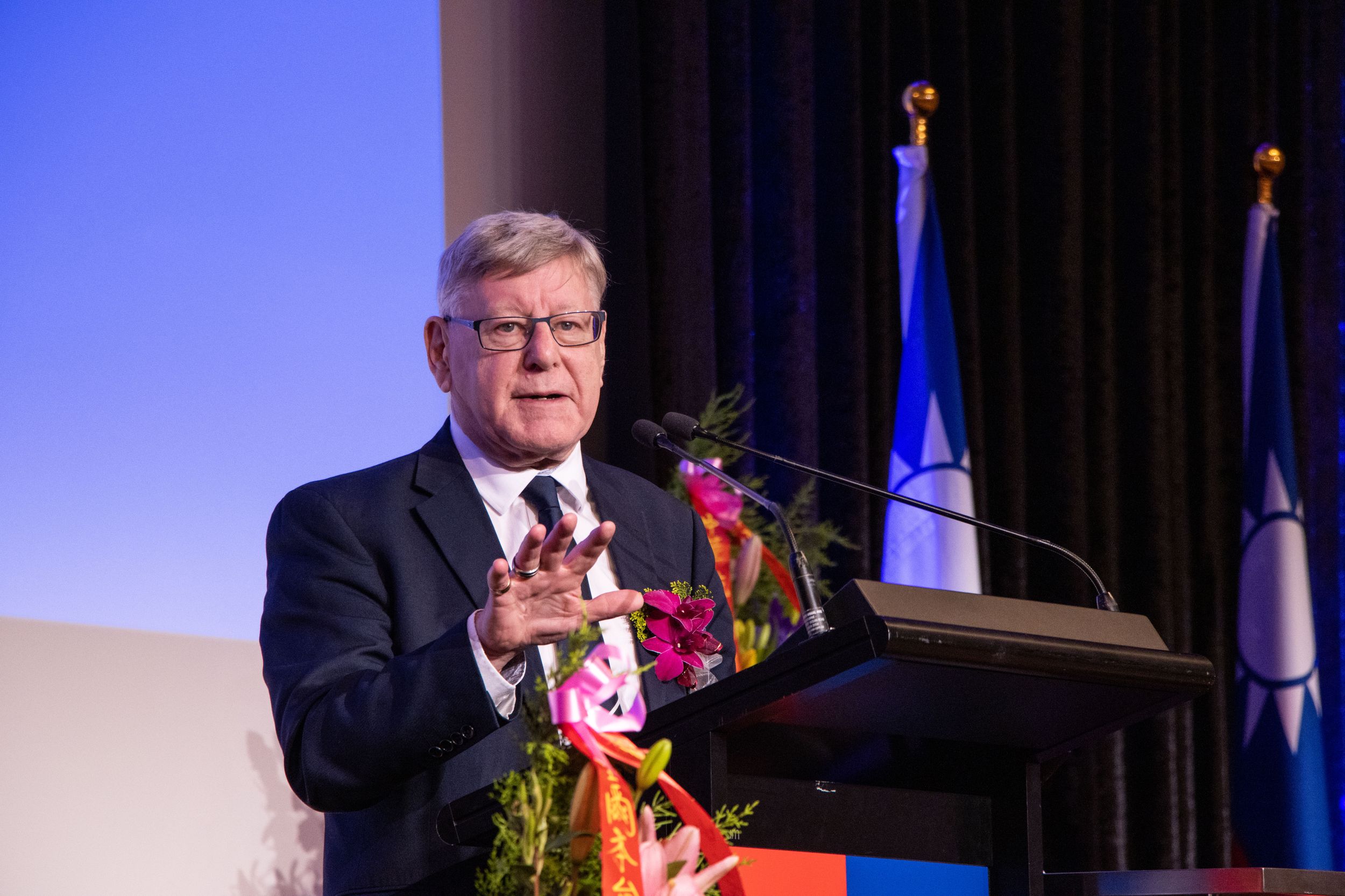 Speaker gestures while presenting at decorated podium with Taiwan flag in background.