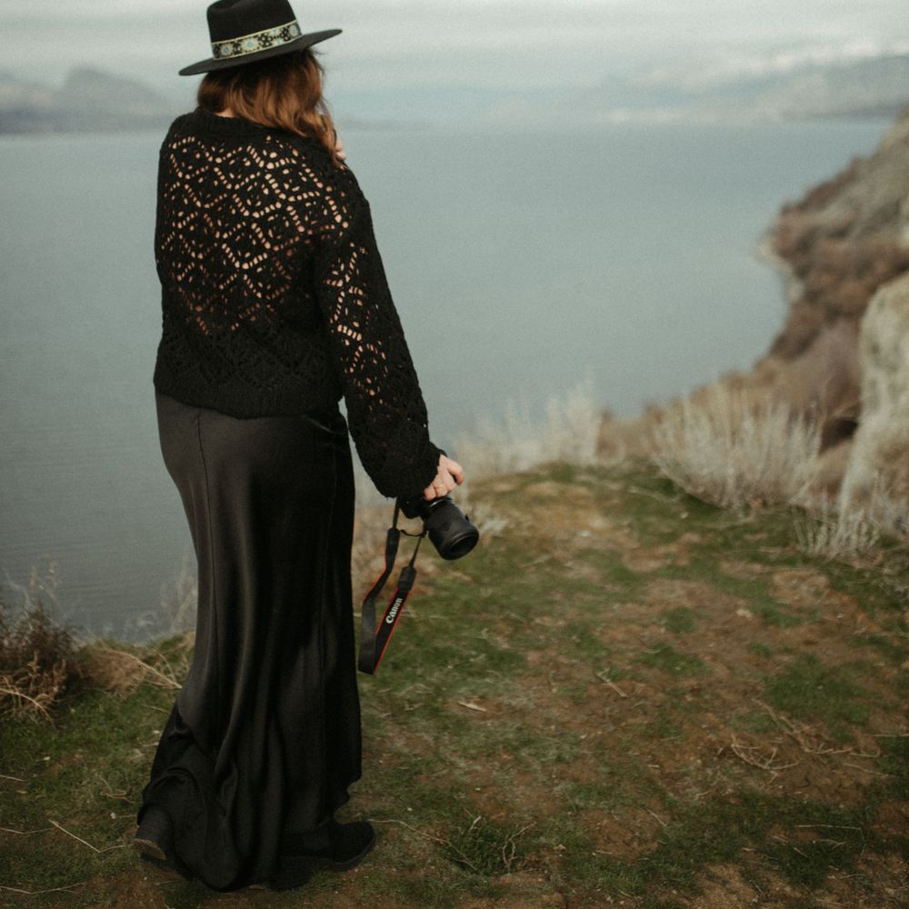 Woman in black lace top and skirt holding camera overlooking a lake from a cliff.