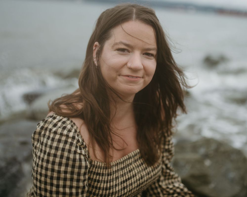 Young woman with brown hair smiling softly while sitting near rocky shoreline on overcast day.
