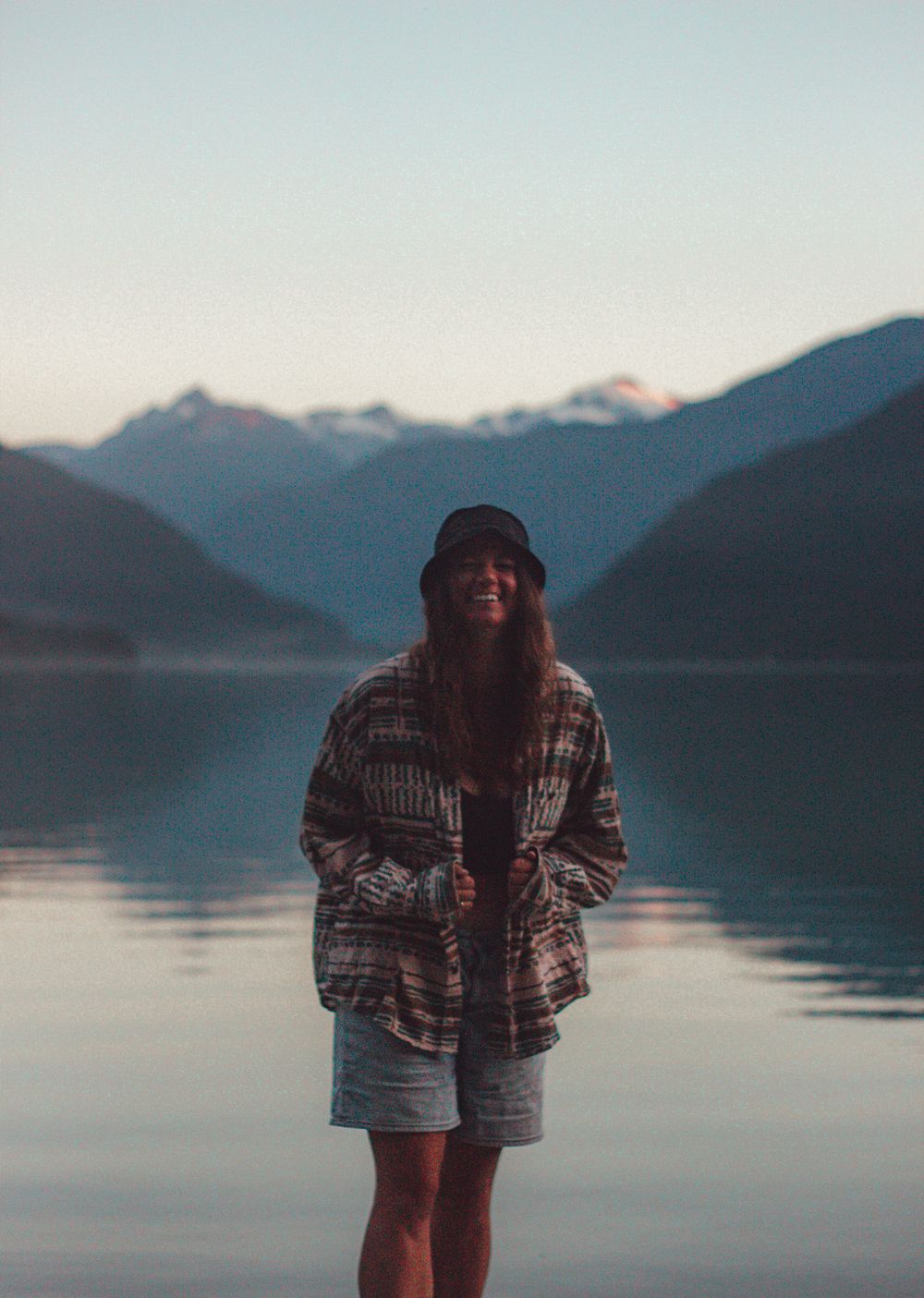 Woman smiling standing in front of a mountain lake at dusk wearing a patterned jacket.