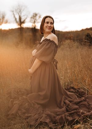 Person in brown dress poses gracefully in golden sunlit field during sunset creating ethereal autumn atmosphere.
