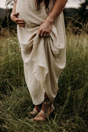 Person wearing flowing cream dress and vintage boots stands in tall grass field at sunset.